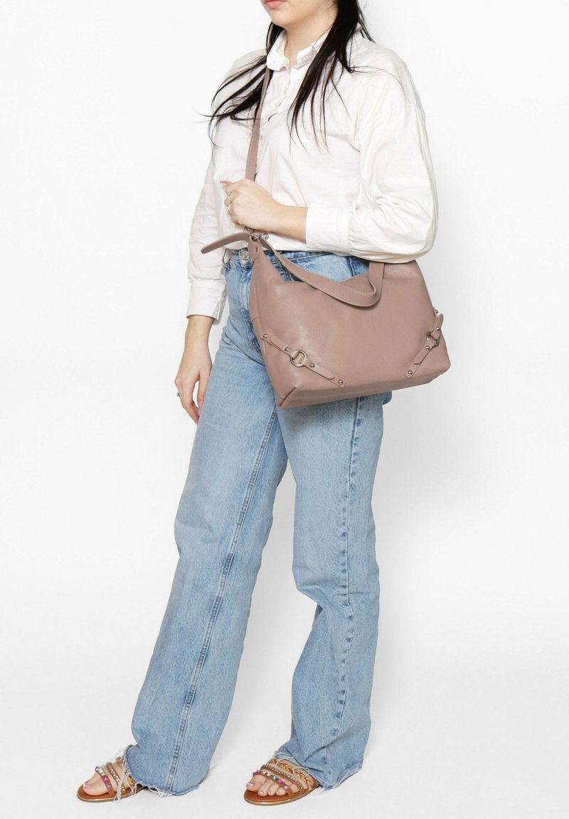Bruine leren schoudertas met een gestructureerde afwerking, voorzien van zilveren hardware-accenten en een gebogen ontwerp, gedragen door een persoon in een lichte outfit.