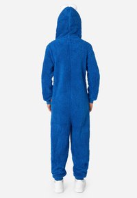 Modra flisasta pižama z mehko teksturo, ki ima kapuco in bele pompone na vrhu. Rokavi in hlače so zaključeni in prilegajoče za udoben fit.