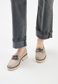 Mocassini in pelle beige con una catena decorativa sulla parte anteriore, caratterizzati da una suola spessa e una finitura testurizzata, abbinati a jeans scuri arrotolati.