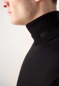 Pulover negru cu guler înalt, realizat dintr-un material tricotat neted, având un guler cu model strapunt cu un logo discret "HUGO" pe gât.