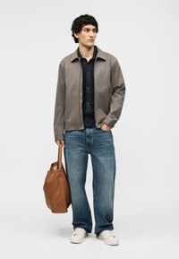 Άνδρας που φορά γκρι μπουφάν, μαύρο πουκάμισο, μπλε τζιν, λευκά αθλητικά παπούτσια, κρατώντας καφέ δερμάτινη τσάντα, στέκεται μπροστά σε απλό φόντο.