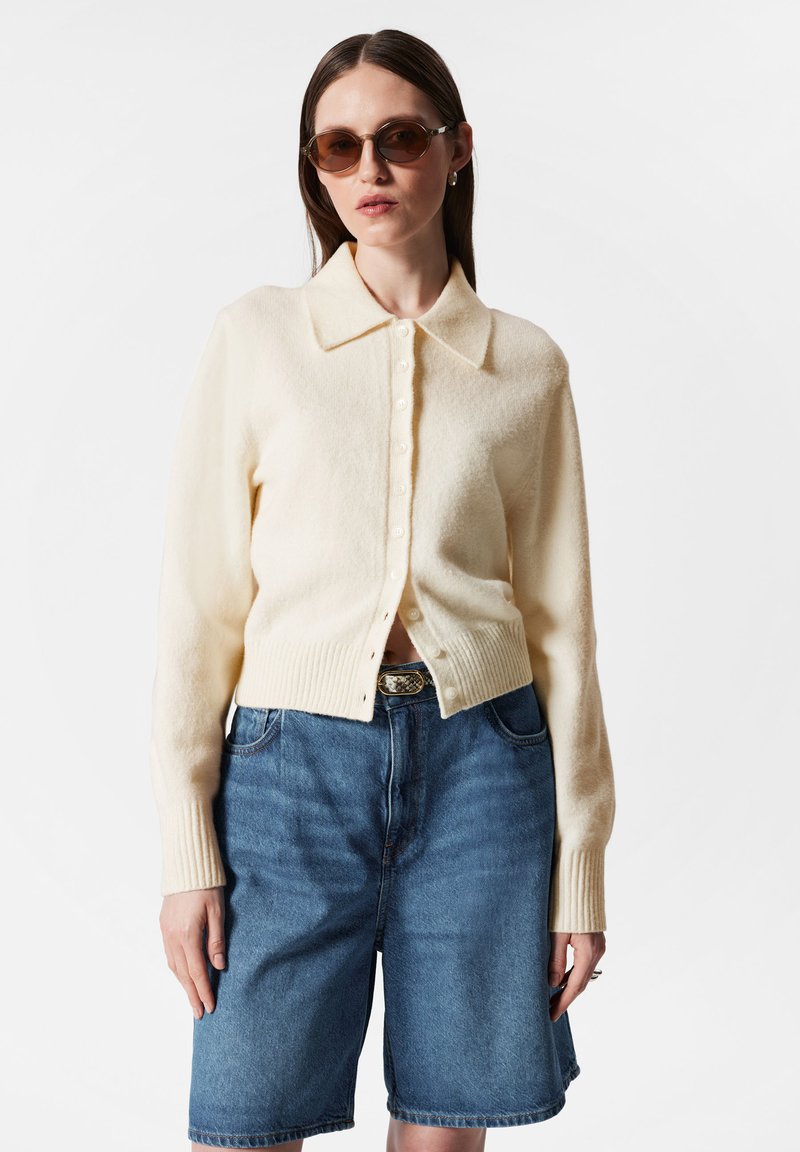 Cremfarbener Cardigan mit spitzem Kragen, gerippten Bündchen und Knopfverschluss, kombiniert mit mid-blauen Denim-Shorts. Glatte Textur, lässige Passform.