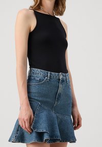 Vrouw in een mouwloos zwart topje, gestoken in een blauwe denim rok met een gerimpelde zoom, staande tegen een effen lichte achtergrond.