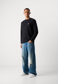 Mand iført briller, sort sweatshirt, løse, slidte blå jeans, hvide sneakers, stående med den ene hånd i lommen mod en ensfarvet baggrund.