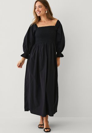 Kvinde smilende, iført lang sort kjole med puffede 3/4 ærmer og firkantet udskæring, kombineret med sorte åbne hæle.