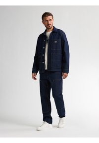 Mørkeblå denimjakke og bukser, med knaplukninger, lomme detaljer og kontrastsyninger, parret med hvide sneakers.