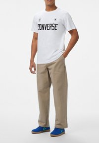 Camiseta blanca de manga corta con impresión negra "CONVERSE", pantalones beiges y zapatillas azules con acentos verdes. Conjunto casual con un diseño minimalista.