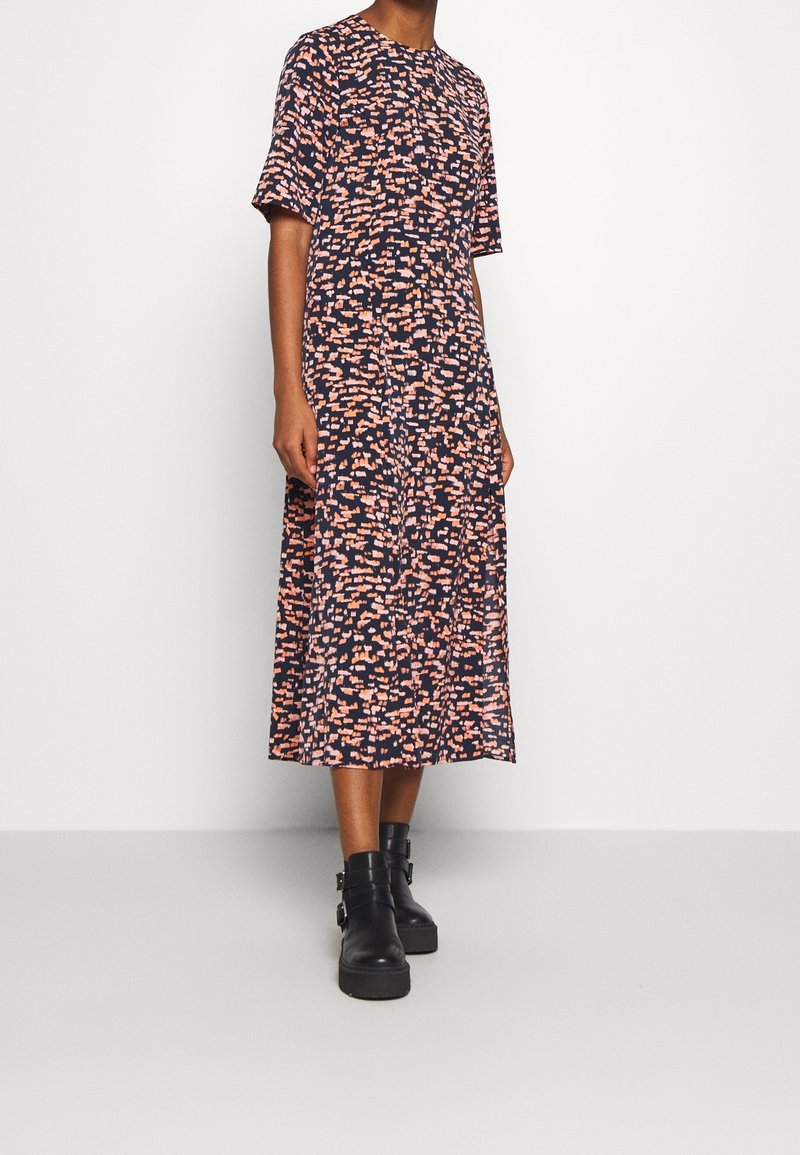 Femme portant une robe midi à manches courtes avec un motif abstrait rose et noir, associée à des bottines noires à boucles.