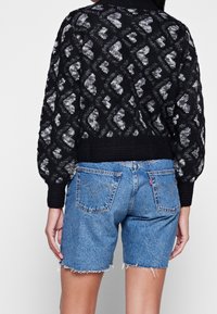 Nainen pukeutuneena musta-harmaaseen kirjavapintaiseen neulepaitaan ja sinisiin farkkushortseihin, joissa on risareunat, kuvattuna takaa.