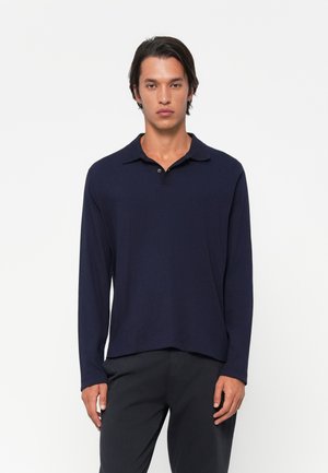 Man wearing a navy long-sleeve polo shirt and dark pants, standing against a plain white background with a neutral expression.