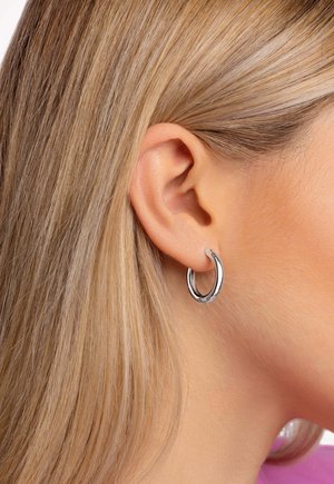Gros plan de l'oreille d'une femme blonde portant une petite boucle d'oreille argentée brillante sur un fond clair.