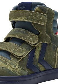 Gröna mockaskor med hög klack som har blå accenter, två kardborreband, en strukturerad kamouflagemönster och en vadderad krage.