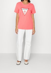 Koraleowy t-shirt z trójkątną białą i czerwoną grafiką z napisem "GUESS" i znakiem zapytania. Połączony z białymi spodniami i beżowymi szpilkami.