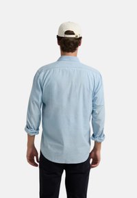 Camisa de botón azul claro hecha de una tela suave, con mangas enrolladas y un dobladillo curvo, llevada con una gorra blanca.