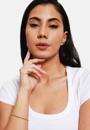 Bracciale in oro su un polso, persona con pelle di media abbronzatura e capelli scuri, che indossa una maglietta bianca a coste, con trucco e unghie in toni neutri.