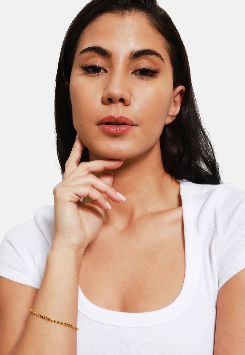 Bracciale in oro su un polso, persona con pelle di media abbronzatura e capelli scuri, che indossa una maglietta bianca a coste, con trucco e unghie in toni neutri.