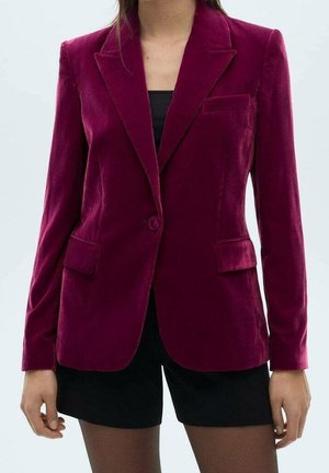 Femme portant un blazer en velours rouge profond ajusté avec des revers crantés et des poches à rabat, sur un short et un haut noirs.
