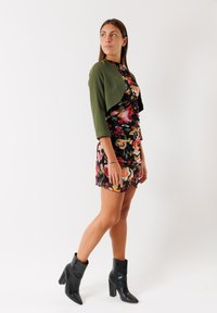 Abito floreale con giacca verde corta, lunghezza corta, design arricciato, sfondo nero con petali colorati, abbinato a stivaletti neri con tacco.