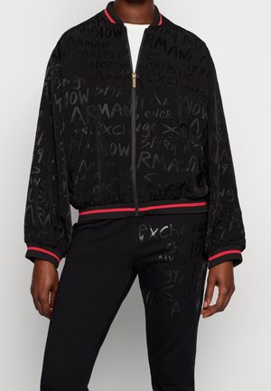 Chaqueta con cremallera y pantalones negros con ribetes rojos, con palabras en estilo graffiti como "Armani" y "Exchange" en diferentes tipografías y tonalidades.