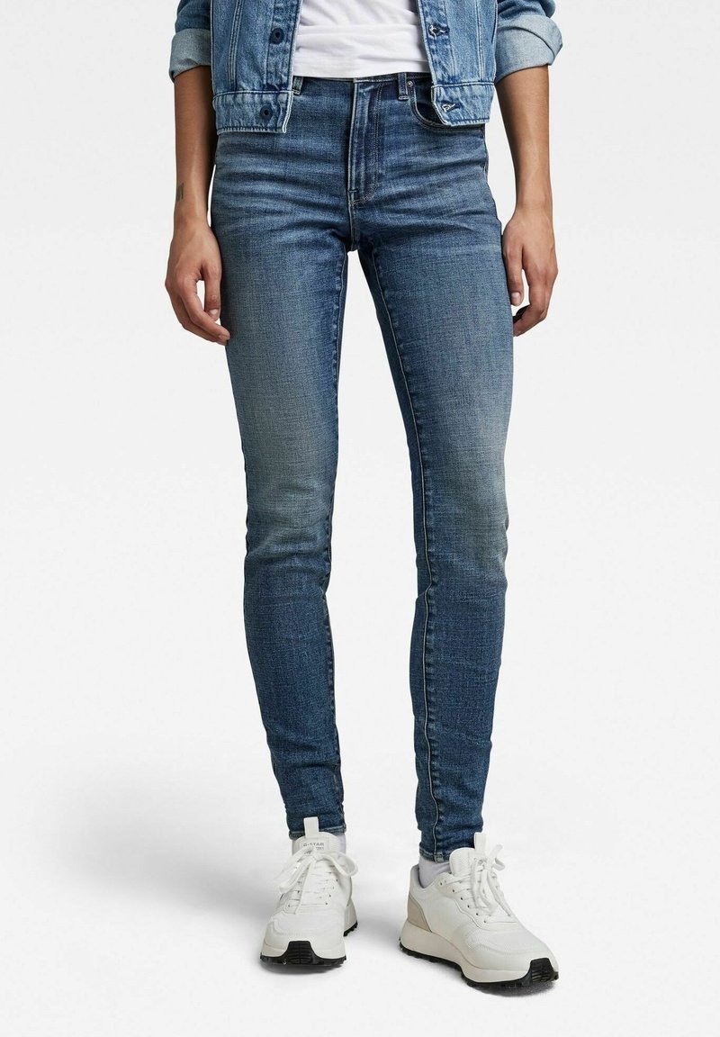 Personne portant un jean bleu ajusté, des baskets blanches et une veste en denim aux manches retroussées, debout devant un fond blanc uni.