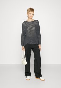Maglione grigio a maglia con maniche larghe e una vestibilità ampia, abbinato a pantaloni neri e sneakers bianche. Tiene una piccola borsa bianca.