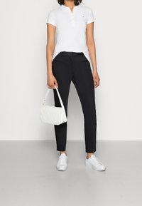 Biała koszulka polo z guzikami, czarne spodnie slim-fit i białe sneakersy. Trzyma białą pikowaną torebkę na ramię.