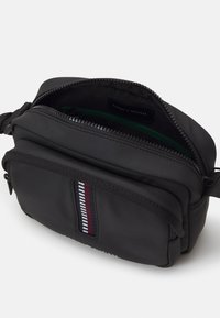 Črn nylon torbica za čez ramo z zadrgo, ki ima zeleno notranjo oblogo in črtast vzorec v rdeči, beli in modri barvi.