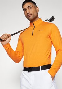 Oranžna srajca z dolgimi rokavi in zadrgo na ovratniku, z diskretno oznako "Calvin Klein". Sestavljena z belimi hlačami in črnim pasom.
