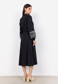 Vestido negro con mangas abullonadas y bordado floral blanco en los puños. Silueta en A, hasta la rodilla, combinado con sandalias de plataforma beige.
