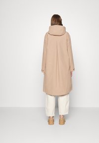 Beiger, knielanger Mantel mit Kapuze, flacher Rückenfalte und knöpfbaren Manschetten. Kombiniert mit hellen, locker sitzenden Hosen und beigen Schuhen.