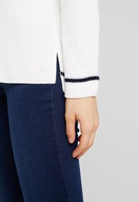 Pull en maille blanche avec un motif texturé et des rayures bleu foncé aux poignets des manches, associé à un jean bleu foncé.