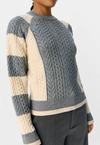 Donna che indossa un maglione grigio e avorio lavorato a maglia con maniche a righe e orlo a costine, abbinato a pantaloni grigio scuro.