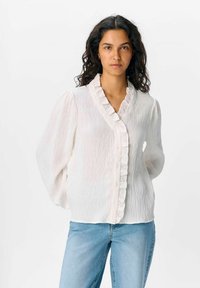 Blusa bianca con texture, colletto arricciato e chiusura con bottoni, dotata di maniche lunghe. Abbinala a jeans azzurri chiari.