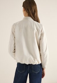 Femme aux longs cheveux bruns portant une veste bomber beige clair et un jean bleu foncé, vue de dos sur un fond neutre.