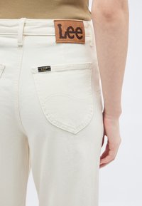 Personne portant un jean écru avec une poche arrière et un patch en cuir marron de la marque Lee sur la ceinture, côté visible du bras droit.