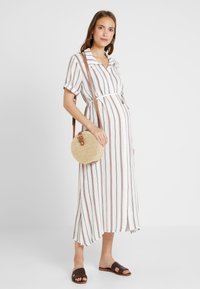Vestido midi listrado em branco com listras castanhas e ferrugentas, com fecho de botões à frente e cintura cingida. Acompanhado de uma bolsa redonda de palha e sandálias escuras.