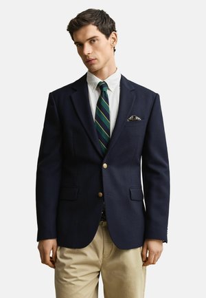 Mann trägt marineblauen Blazer mit goldenen Knöpfen, weißes Hemd, grün gestreifte Krawatte, gemustertes Einstecktuch und beige Hose vor weißem Hintergrund.