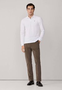 Valkoinen pitkähihainen poolopaita, jossa on nappilista ja logo, yhdistettynä ruskeisiin kapealahkeisiin housuihin ja mustiin kenkiin. Kuvioitu kangas sopii rentoon pukeutumiseen.