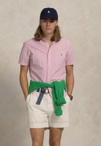 Růžová kostkovaná košile s krátkým rukávem, bílé šortky se zeleným svetrem uvázaným v pase a tmavě modrá kšiltovka s logem na přední straně.