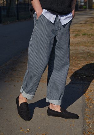 Gestreepte blauwe en witte losse broek met opgerolde pijpen, gecombineerd met zwarte suède loafers. Een wit overhemd met kraag onder een donkere trui.