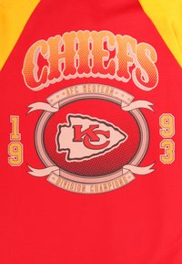 Punainen paita keltaisilla hihoilla, jossa on "CHIEFS"-teksti, AFC Western -logo ja "DIVISION CHAMPIONS" -banneri erottuvilla väreillä ja kuvioilla.