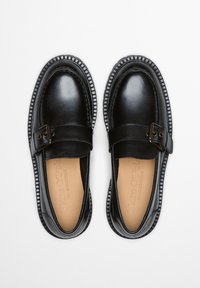 Czarne mokasyny ze skóry o gładkim wykończeniu, z paskiem na klamrę i białą, teksturowaną gumową podeszwą. Wnętrze wyściełane jasnobrązową podszewką z brandingiem.