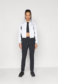 Balta sporta jakas ar melniem akcentiem, saskaņota ar melnu crop top un melnām biksēm. Uz jakas un biksēm ir logo; melni sporta apavi.