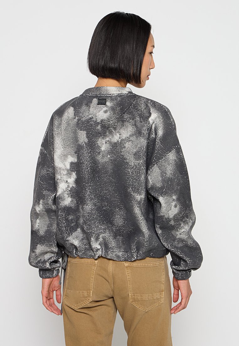 Grauer Pullover mit Spritzmuster, gerippten Bündchen und Saum, weicher Textur, lockerer Passform und dezentem Branding am hinteren Halsausschnitt.