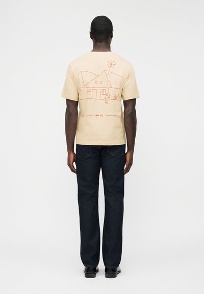 Άνδρας που φοράει μπεζ μπλουζάκι με κόκκινο σχέδιο γραμμής που απεικονίζει σπίτι, δέντρο, ποδήλατο και άνθρωπο στην πλάτη, συνδυασμένο με σκούρο τζιν και μαύρα παπούτσια.