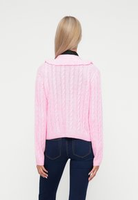 Ροζ πουλόβερ με πλέξιμο και κοντό σχέδιο, διαθέτει γιακά. Μαλακή υφή και ριμπ τελείωμα. Φοριέται με σκούρα μπλε τζιν, απεικονίζοντας την πίσω όψη.