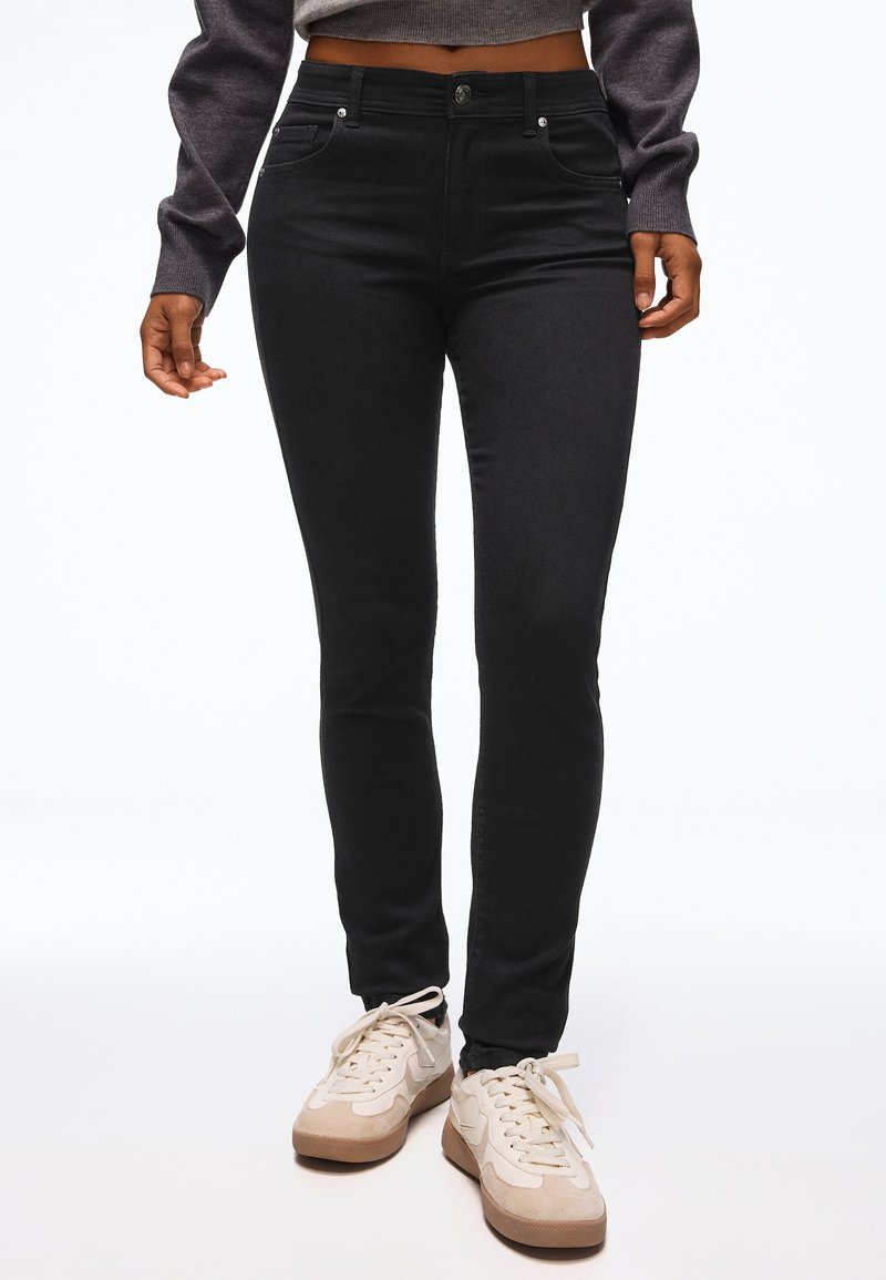 Personne portant un jean skinny noir, des baskets beige et blanches, et un pull gris court, debout devant un fond blanc uni.