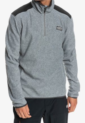 Gray fleece pullover with a high collar and half-zip, featuring black shoulder accents and a logo patch on the chest. Silky texture.