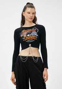 Zwarte geribbelde crop top met lange mouwen, uitsnijding aan de voorkant en een levendig grafisch ontwerp. Voorzien van zilveren kettingen en een smalle tailleband, gecombineerd met zwarte broeken.