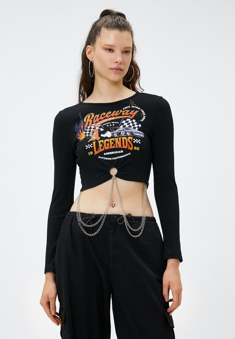 Czarny prążkowany crop top z długimi rękawami, wycięciem z przodu i intensywnym wzorem graficznym. Posiada srebrne łańcuchy i wąski pasek w talii, zestawiony z czarnymi spodniami.